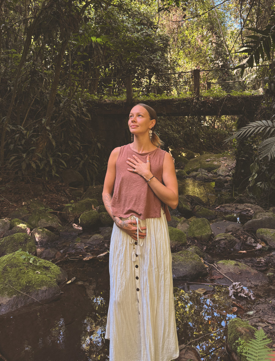 Skye standing beside a jungle stream, one hand resting over her heart, looking up into the light.