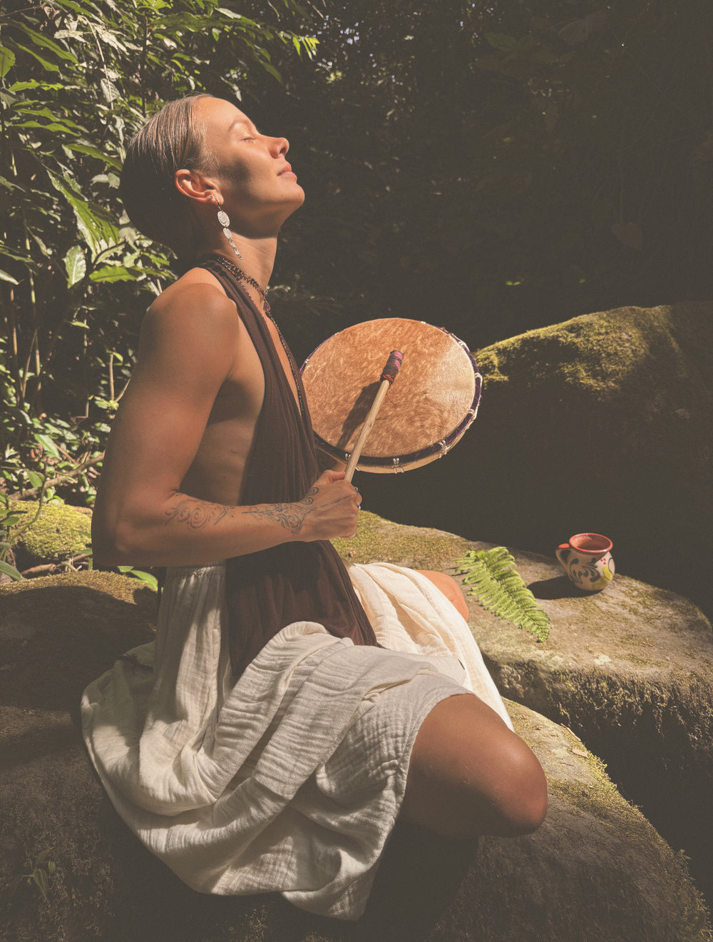 Skye seated on a mossy rock beside the stream, playing her frame drum, face lifted.
