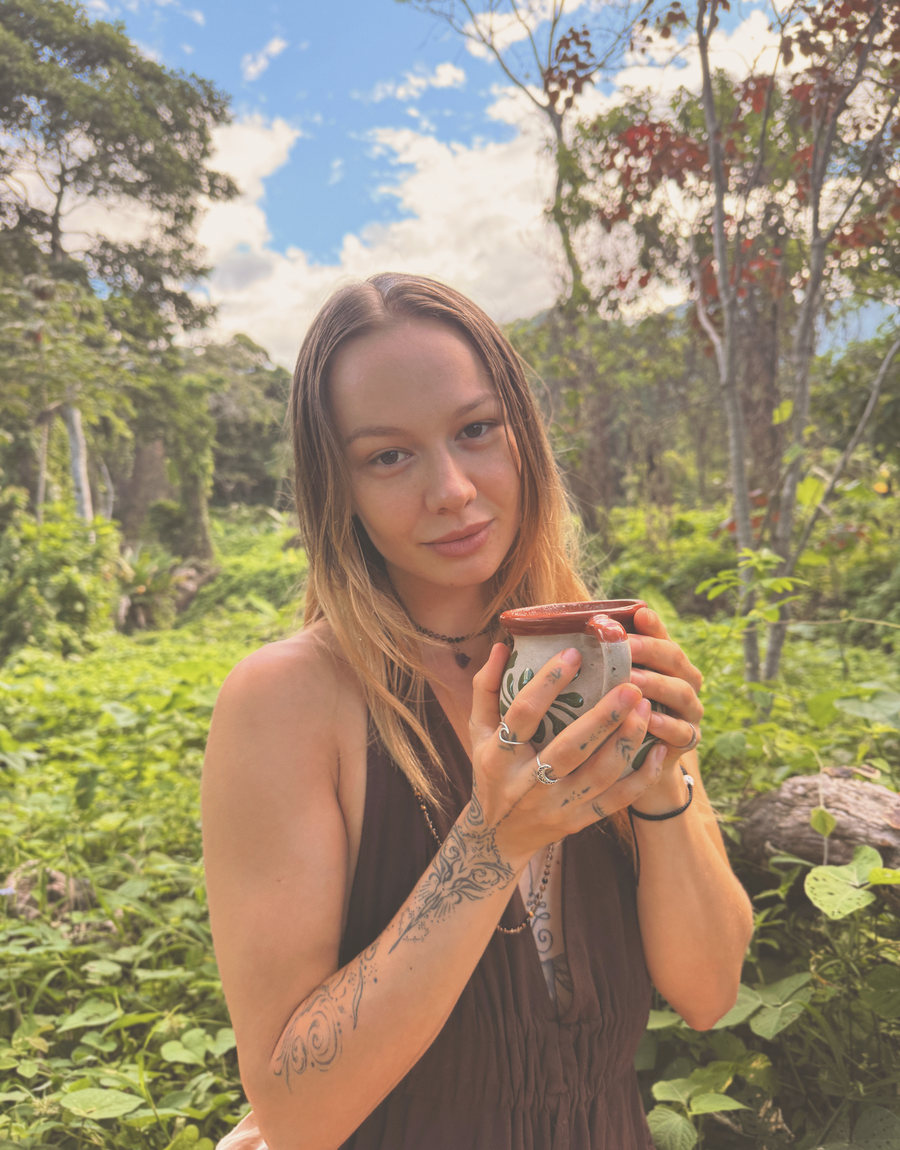 Skye standing in the green, holding a clay cup in both hands, looking at the camera.